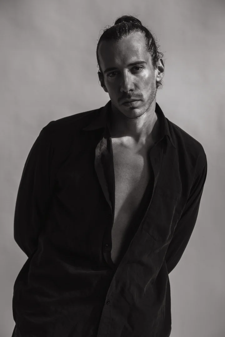 Zeyan Castracane. In an open black shirt, looking directly at the camera. Black-and-white studio portrait.