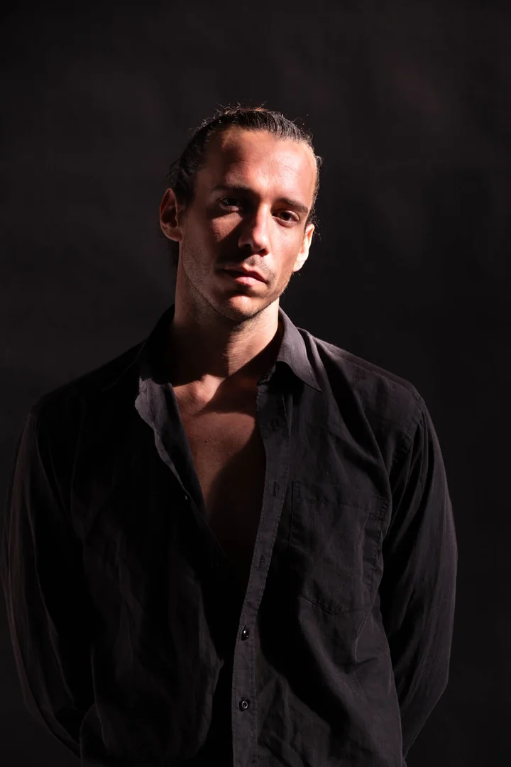 Zeyan Castracane. Standing in a black shirt against a dark backdrop. Colour portrait.