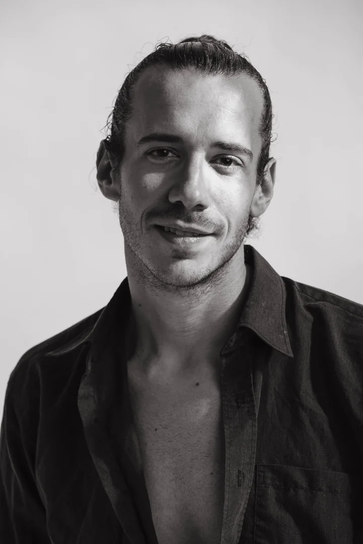 Zeyan Castracane. Smiling in an open black shirt against a light backdrop. Black-and-white portrait.